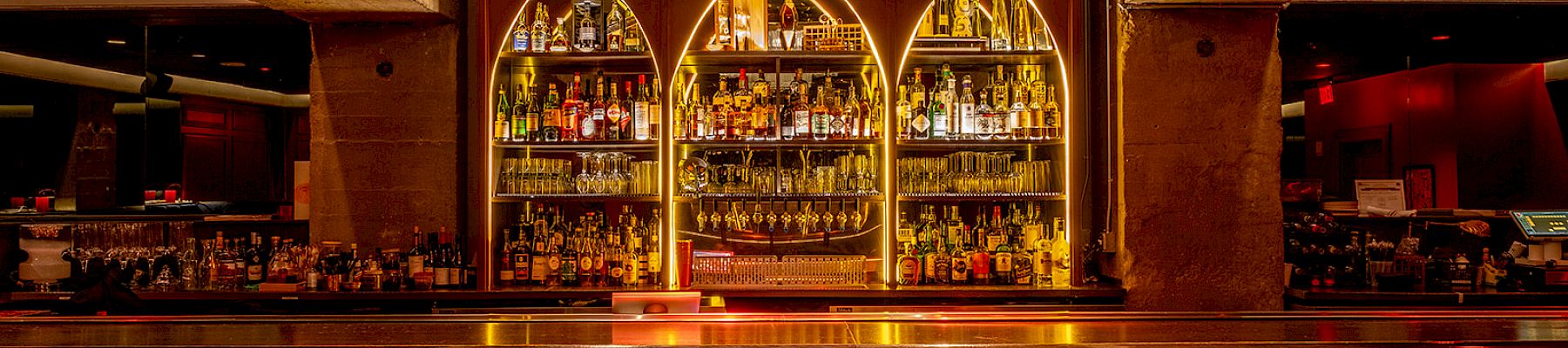 A stylish bar with a row of stools, illuminated shelves with bottles, and a grand chandelier above, creating an elegant atmosphere.