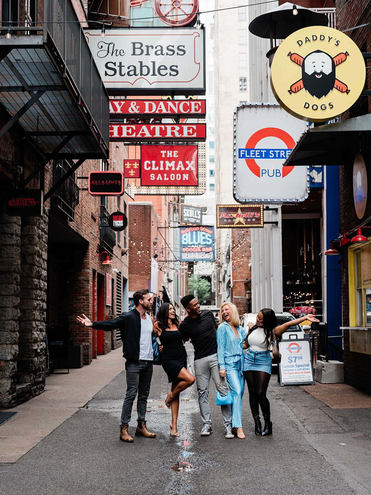 A group of people joyfully pose in an alley with vibrant signs overhead, including “The Brass Stables” and