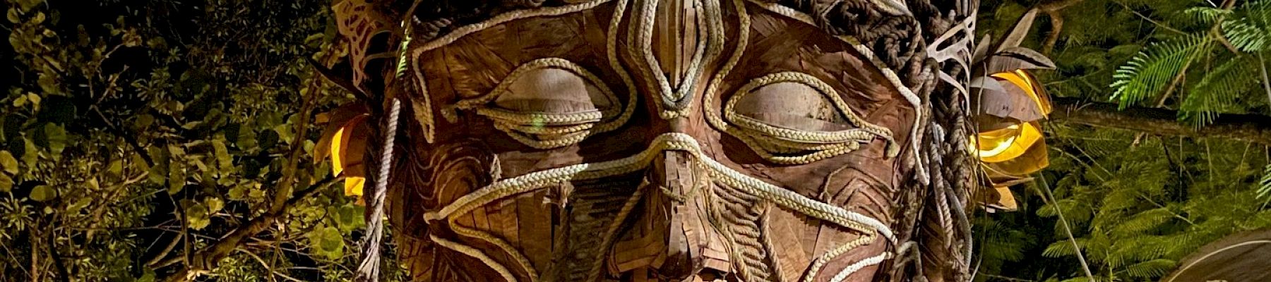 A wooden sculpture of a face with intricate carvings and flowers is surrounded by greenery and dim lighting, blending into the natural setting.
