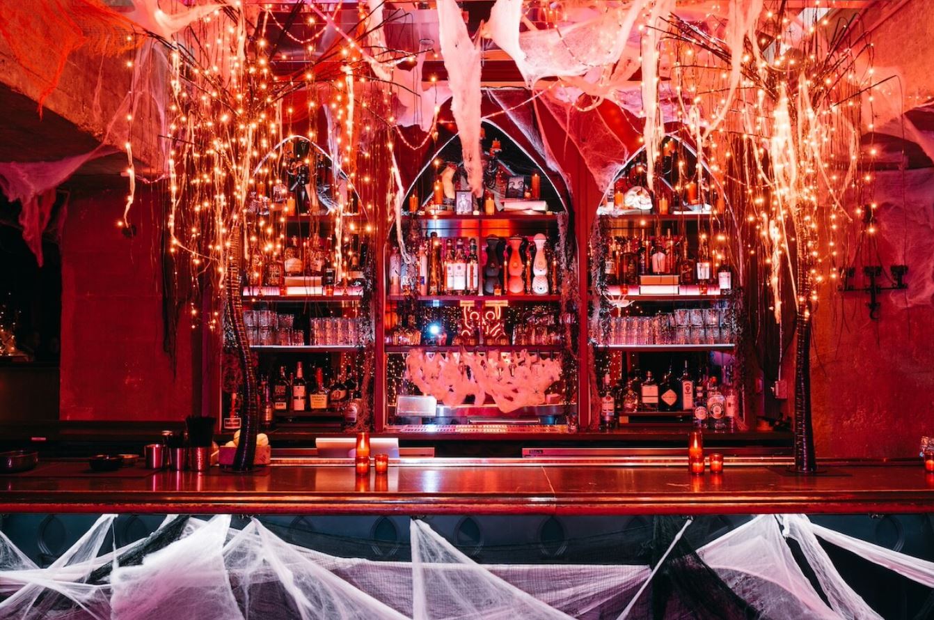 A Halloween-themed bar with cobwebs, dim lighting, and bottles on shelves. The ambiance is spooky and festive.