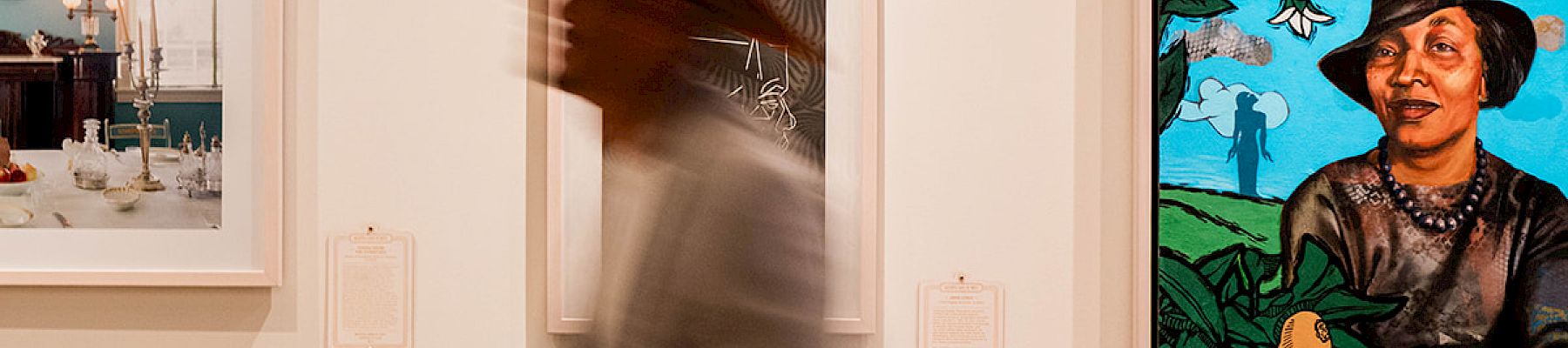 A person in a hat walks past colorful framed art on a gallery wall.