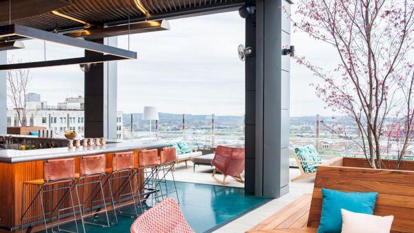 A modern rooftop bar with stylish seating, a wooden counter, and city views. Cozy atmosphere with colorful cushions and trees.