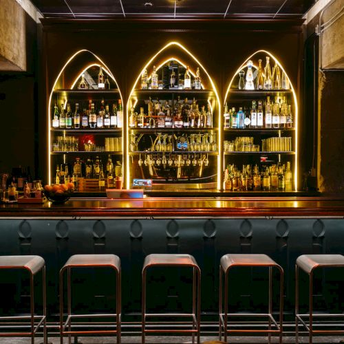A dimly lit bar with stools, a chandelier, and illuminated shelves of bottles.