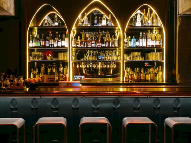 A dimly lit bar with stools, a chandelier, and illuminated shelves of bottles.
