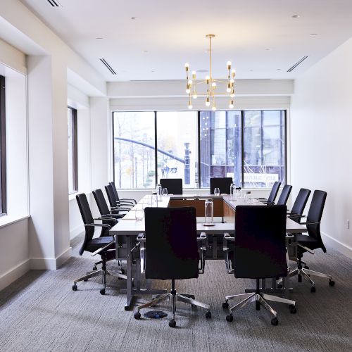 A modern conference room with a long table, office chairs, large windows, and a chandelier, offering a bright office setting.