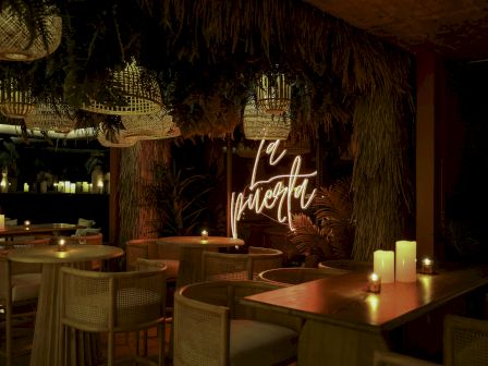 A cozy, dimly lit restaurant with candles on wooden tables, thatched decor, and a neon sign reading 