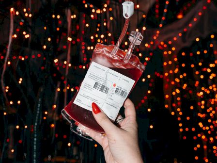 A hand holds a bag resembling a blood bag, possibly a prop, against a background of red and orange lights, creating a spooky atmosphere.