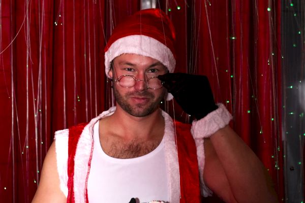 A person dressed in a festive Santa outfit with a sleeveless top and hat, holding a drink in front of a red curtain backdrop.