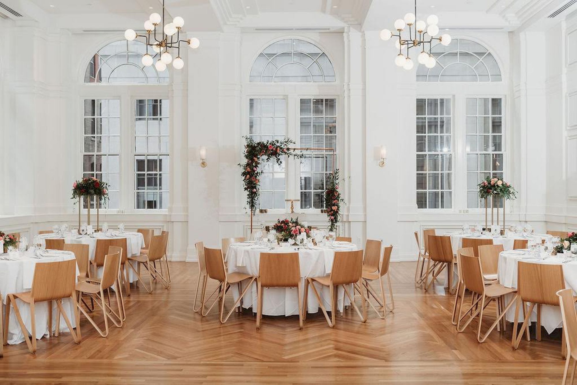A decorated banquet hall with round tables, wooden chairs, floral centerpieces, and elegant chandeliers under high arched windows.