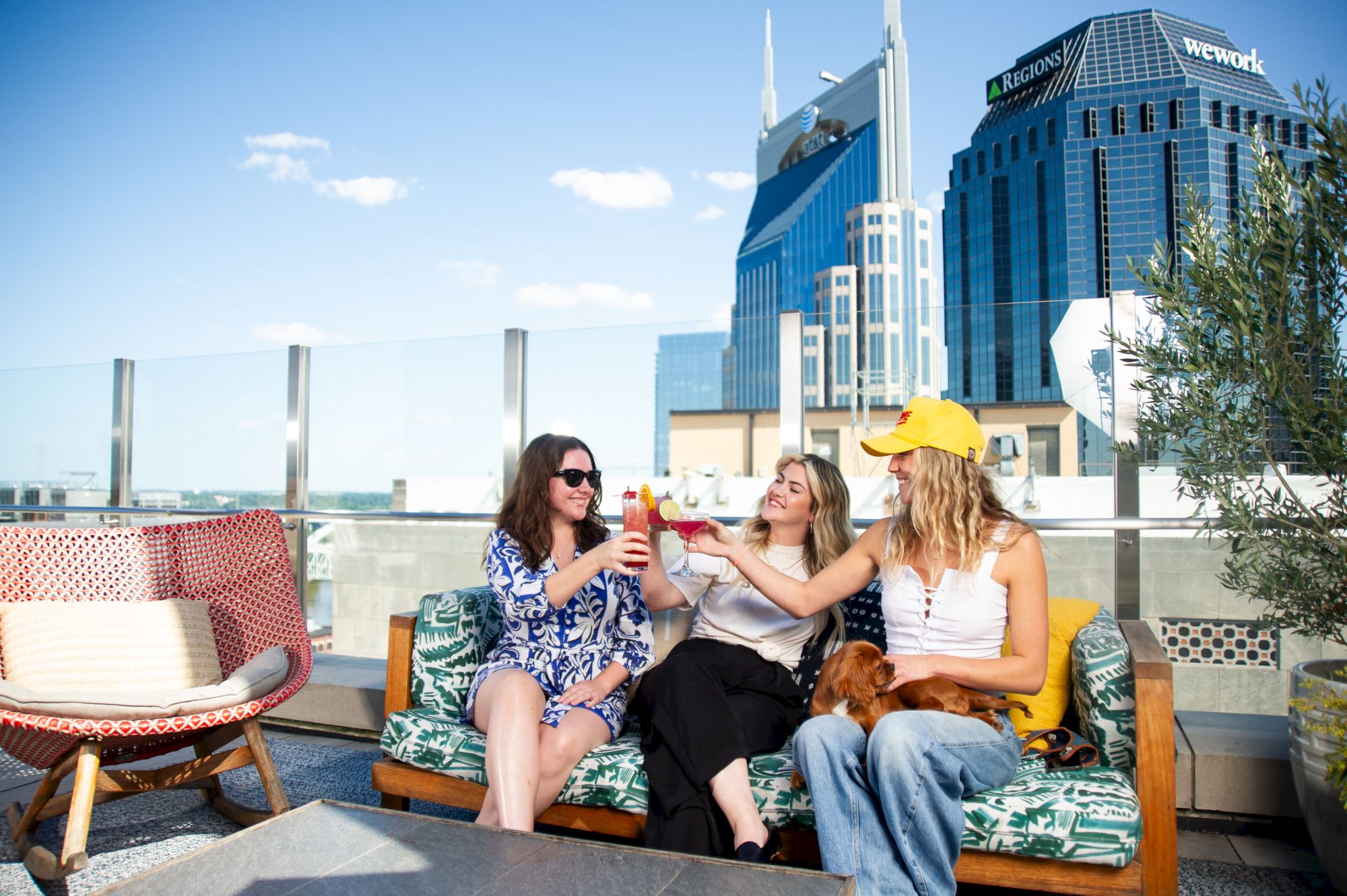 Three people are toasting with drinks on a rooftop patio with city buildings in the background.