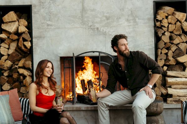 A woman and man sit by a fire, surrounded by stacked firewood and zebra-striped benches, creating a cozy atmosphere.