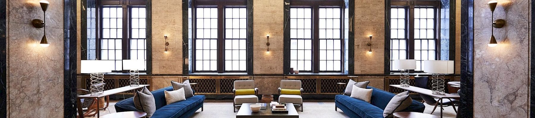 A spacious, elegant room with large arched windows, blue sofas, armchairs, and a coffee table, featuring minimalist decor and soft lighting.