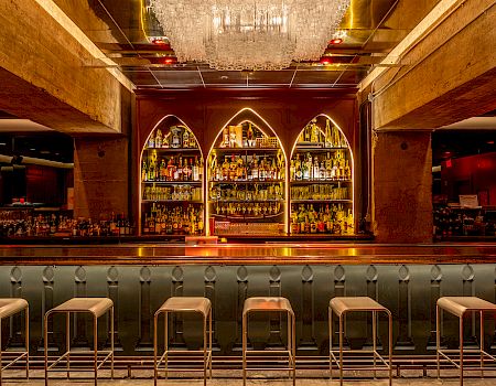 A stylish bar with a row of stools, illuminated shelves with bottles, and a grand chandelier above, creating an elegant atmosphere.