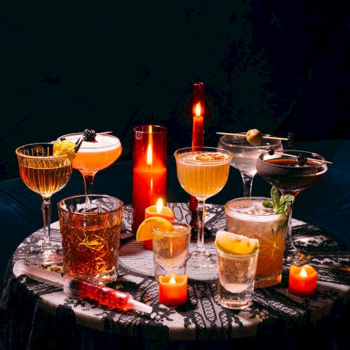 A table with assorted cocktails and candles, including various garnishes like lemons and herbs, creating a moody, ambient setting.