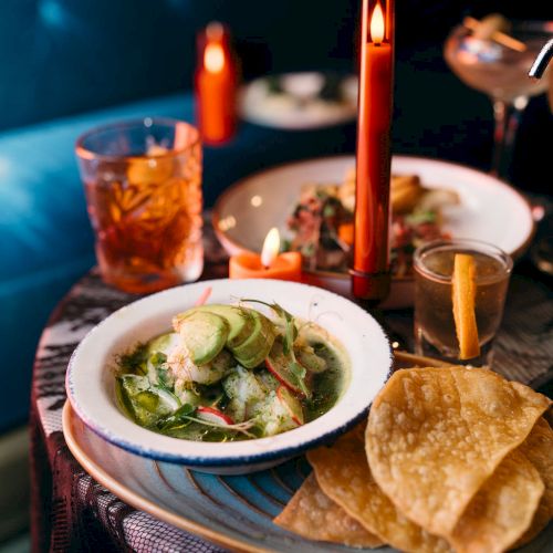 A dimly lit table with a salad, tortilla chips, and drinks, set beside a candle for an intimate dining experience.
