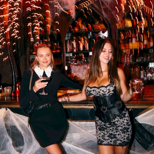 Two women in Halloween costumes posing in a decorated bar with cobwebs and lights.