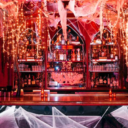 A bar decorated with hanging lights and spider webs, featuring shelves stocked with bottles and glasses, creating a spooky atmosphere.