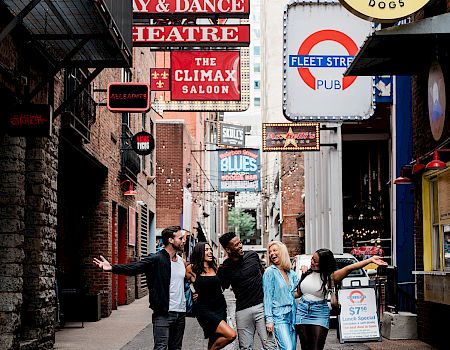 A group of people joyfully pose in an alley with vibrant signs overhead, including “The Brass Stables” and 
