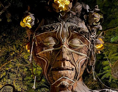 A wooden sculpture of a face with intricate carvings and flowers is surrounded by greenery and dim lighting, blending into the natural setting.