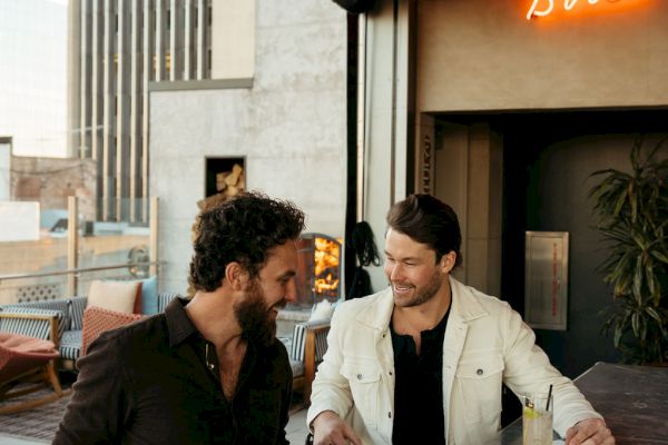 Two people sit at a bar with drinks, talking. A neon sign behind them reads 