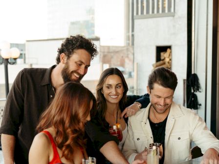 A group of four people socializing at a bar counter, enjoying drinks and looking at a menu or list together, with an outside view.