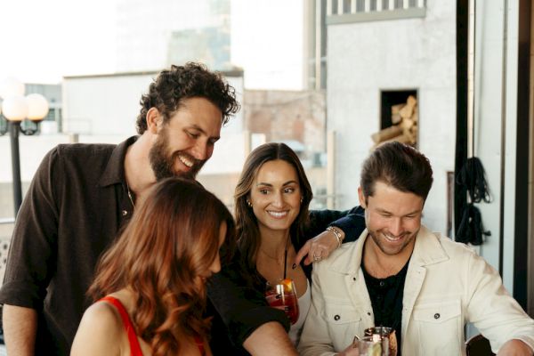 A group of four people socializing at a bar counter, enjoying drinks and looking at a menu or list together, with an outside view.