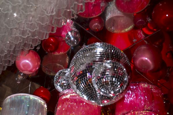 The image shows a collection of colorful, hanging light fixtures, including pink and red lampshades and a shiny silver disco ball.