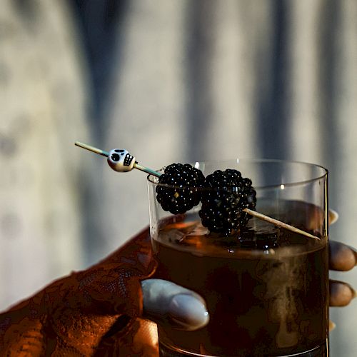 A person is holding a glass of dark-colored drink garnished with blackberries on a skewer, with partial henna design visible on the hand.