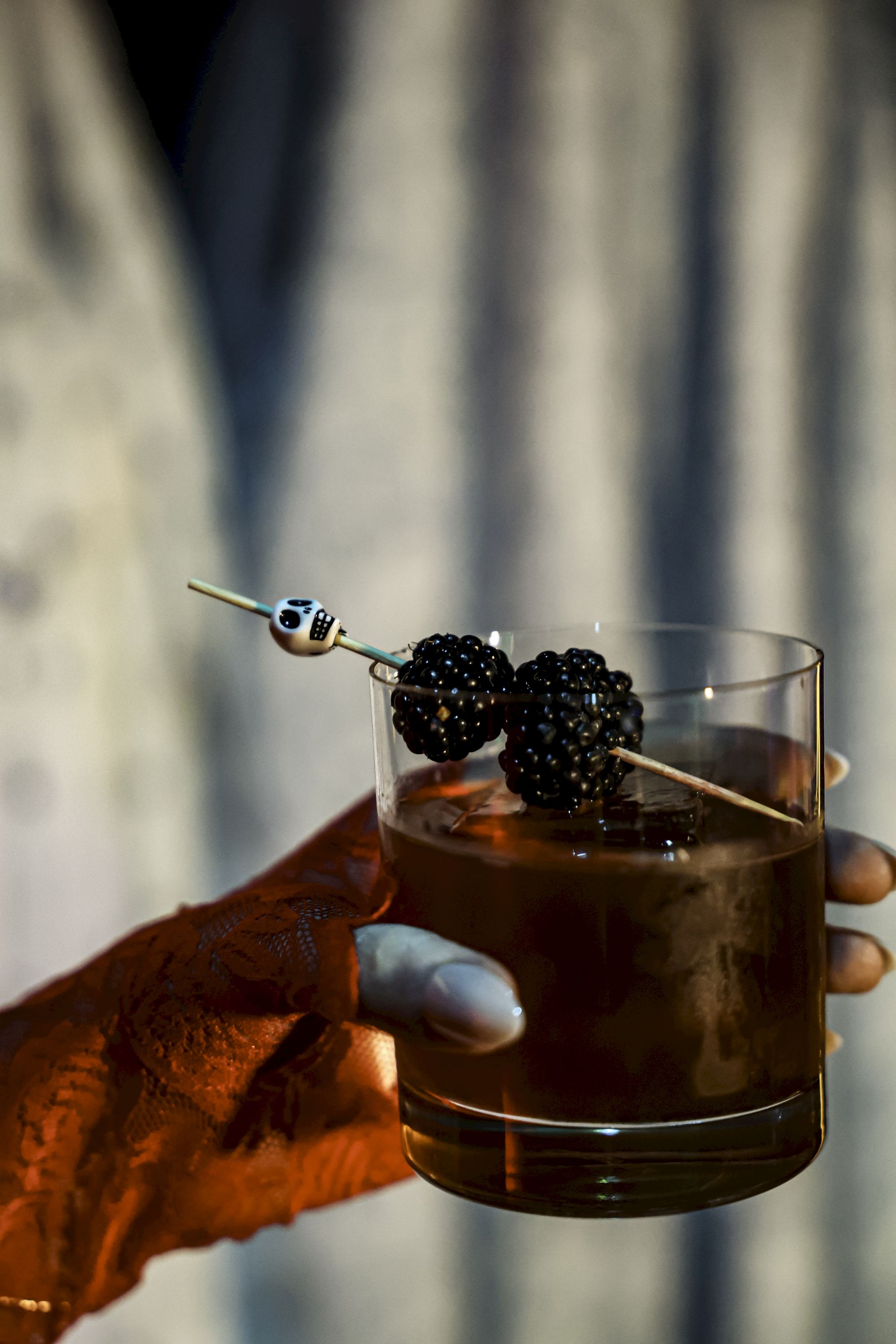 A hand holds a drink with a skewer of blackberries and a decorative skull; intricate henna adorns the hand.
