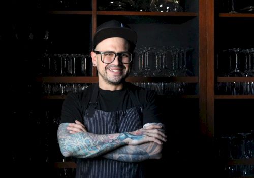 A person with tattoos wearing glasses and a cap, standing with arms crossed, in front of shelves with glassware, smiling.