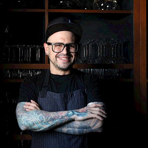A person with tattoos wearing glasses and a cap, standing with arms crossed, in front of shelves with glassware, smiling.
