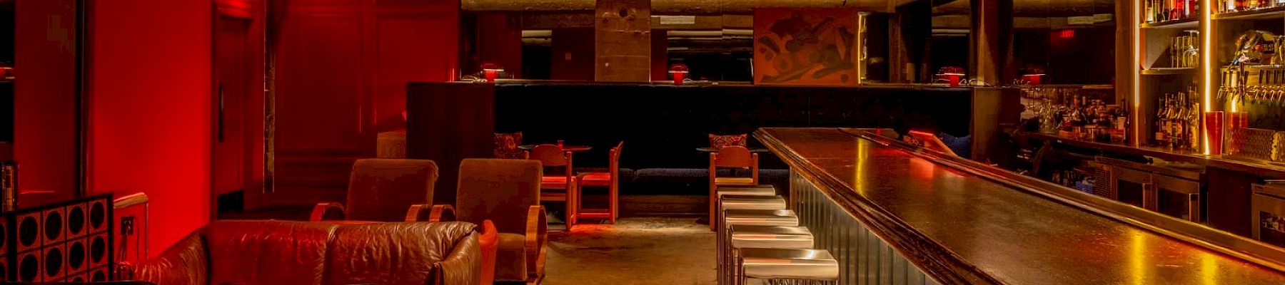 A cozy bar interior with leather seating, a long counter with stools, warm lighting, and a chandelier, creating an inviting ambiance.