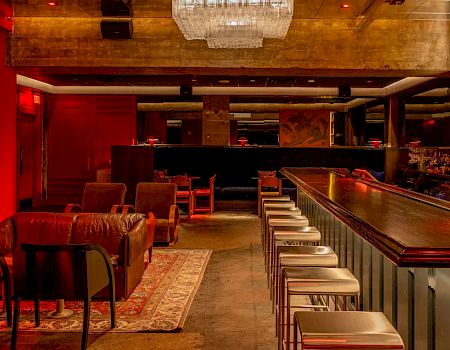 A cozy bar interior with leather seating, a long counter with stools, warm lighting, and a chandelier, creating an inviting ambiance.