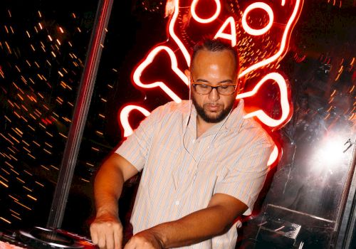 A DJ is playing music with a neon skull and crossbones in the background, surrounded by Halloween-themed decorations.