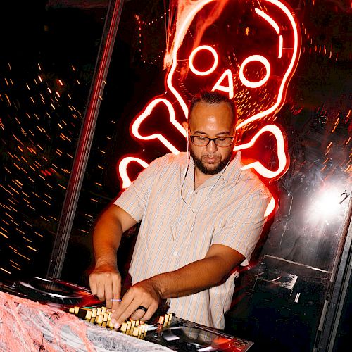 A DJ is playing music with a neon skull and crossbones in the background, surrounded by Halloween-themed decorations.