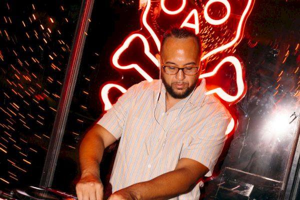 A DJ is performing at a spooky-themed event, with a neon skull and bones sign in the background, and cobweb decorations around.