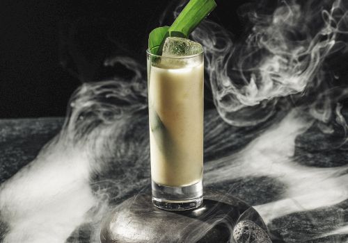 A tall glass filled with a creamy drink, garnished with green leaves, surrounded by swirling smoke on a dark surface.
