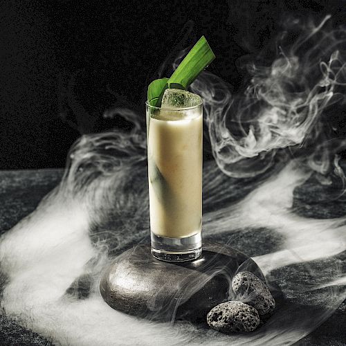 A tall glass filled with a creamy drink, garnished with green leaves, surrounded by swirling smoke on a dark surface.
