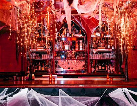 A Halloween-themed bar with cobwebs, dim lighting, and bottles on shelves. The ambiance is spooky and festive.
