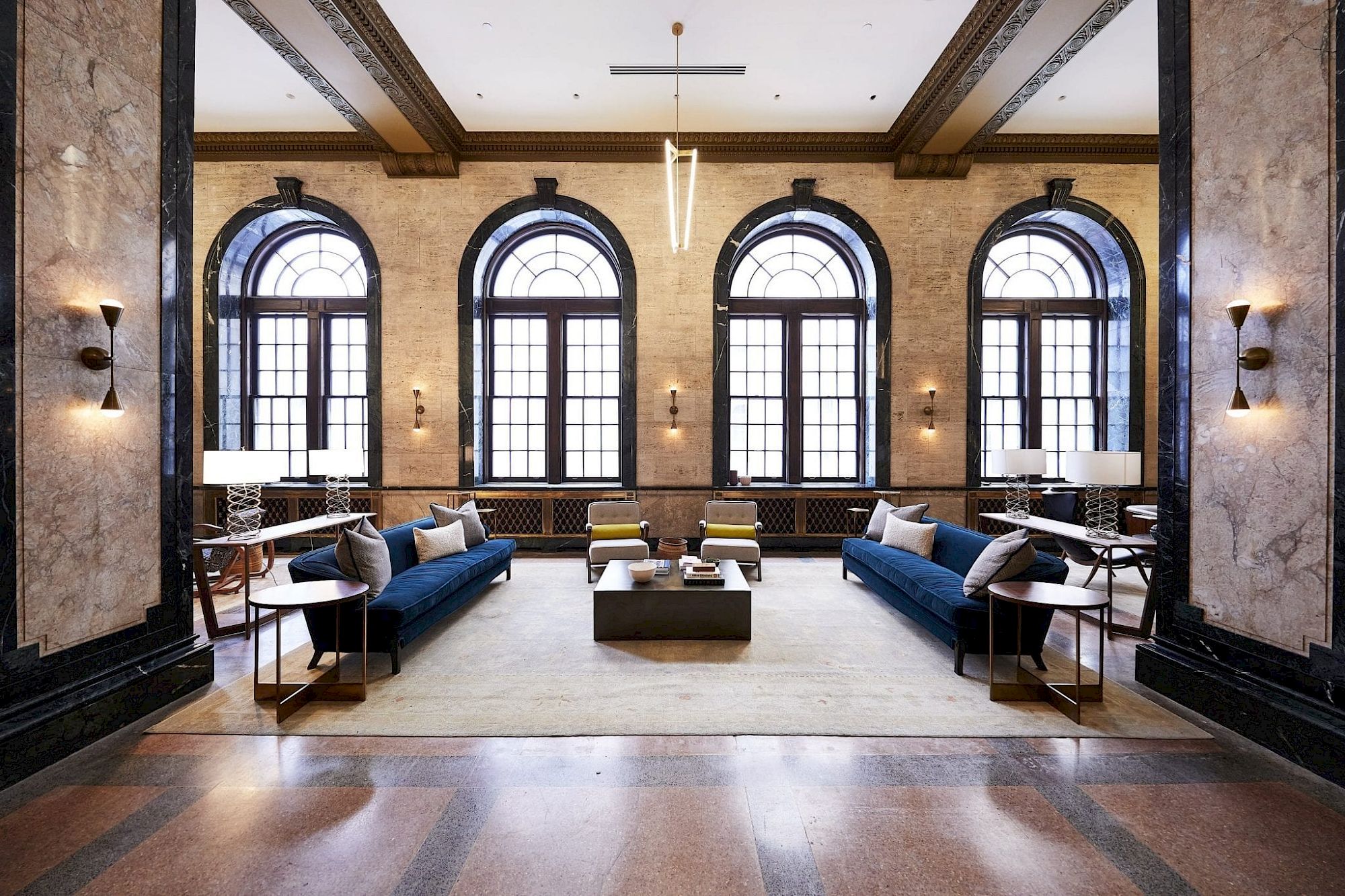 A spacious, elegant room with large arched windows, blue sofas, armchairs, and modern lighting, showcasing sophisticated interior design.