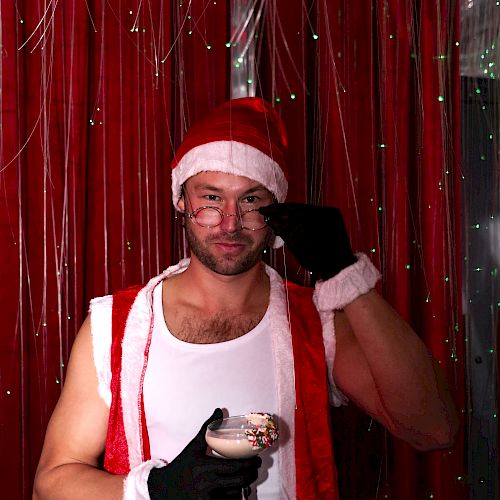 A person in a Santa hat and outfit holds a cocktail, standing in front of a festive backdrop with string lights.