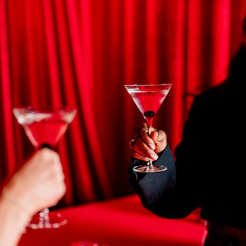 Two people toasting with martini glasses, red curtain background.