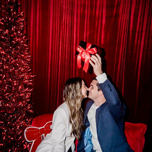A couple is sitting by a red Christmas tree, holding a gift with a red ribbon.