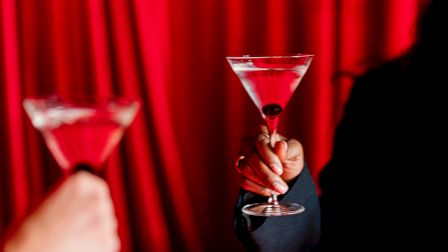 Two people toast with martini glasses against a red curtain backdrop.