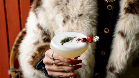 A person in a fur coat holding a cocktail with cranberries and rosemary.