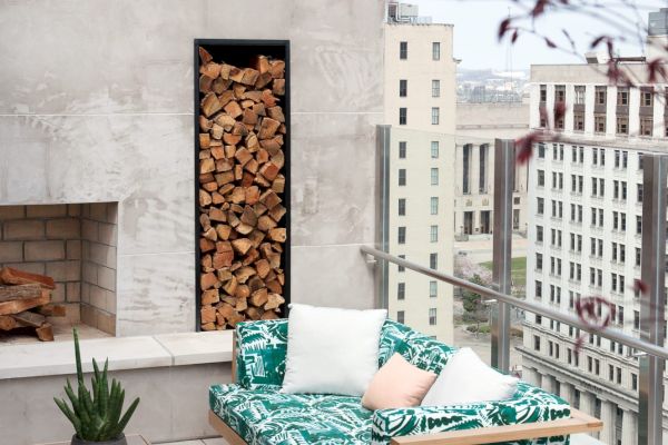 A rooftop patio with outdoor seating, a coffee table, a fireplace with stacked wood, and urban views in the background.