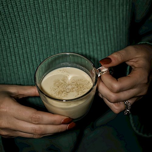 A person wearing a green sweater and gold necklace holds a transparent cup of frothy coffee with red painted nails.