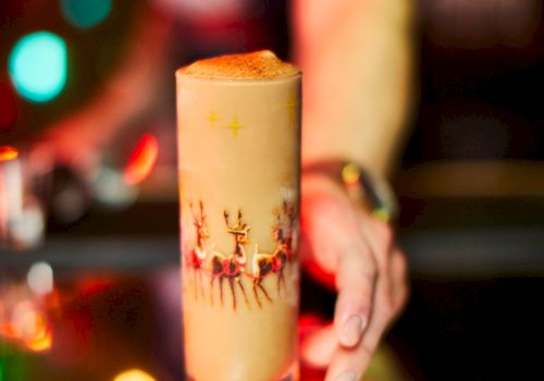 A festive cocktail in a decorated glass on a bar counter with colorful lights in the background and a person reaching towards it.