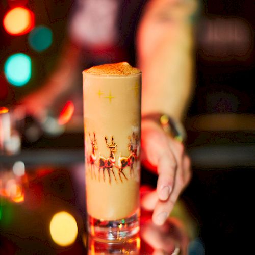 A festive cocktail in a decorated glass on a bar counter with colorful lights in the background and a person reaching towards it.