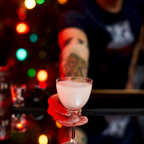 A person is offering a creamy cocktail in a coupe glass, with colorful bokeh lights in the background and a tattooed arm visible.